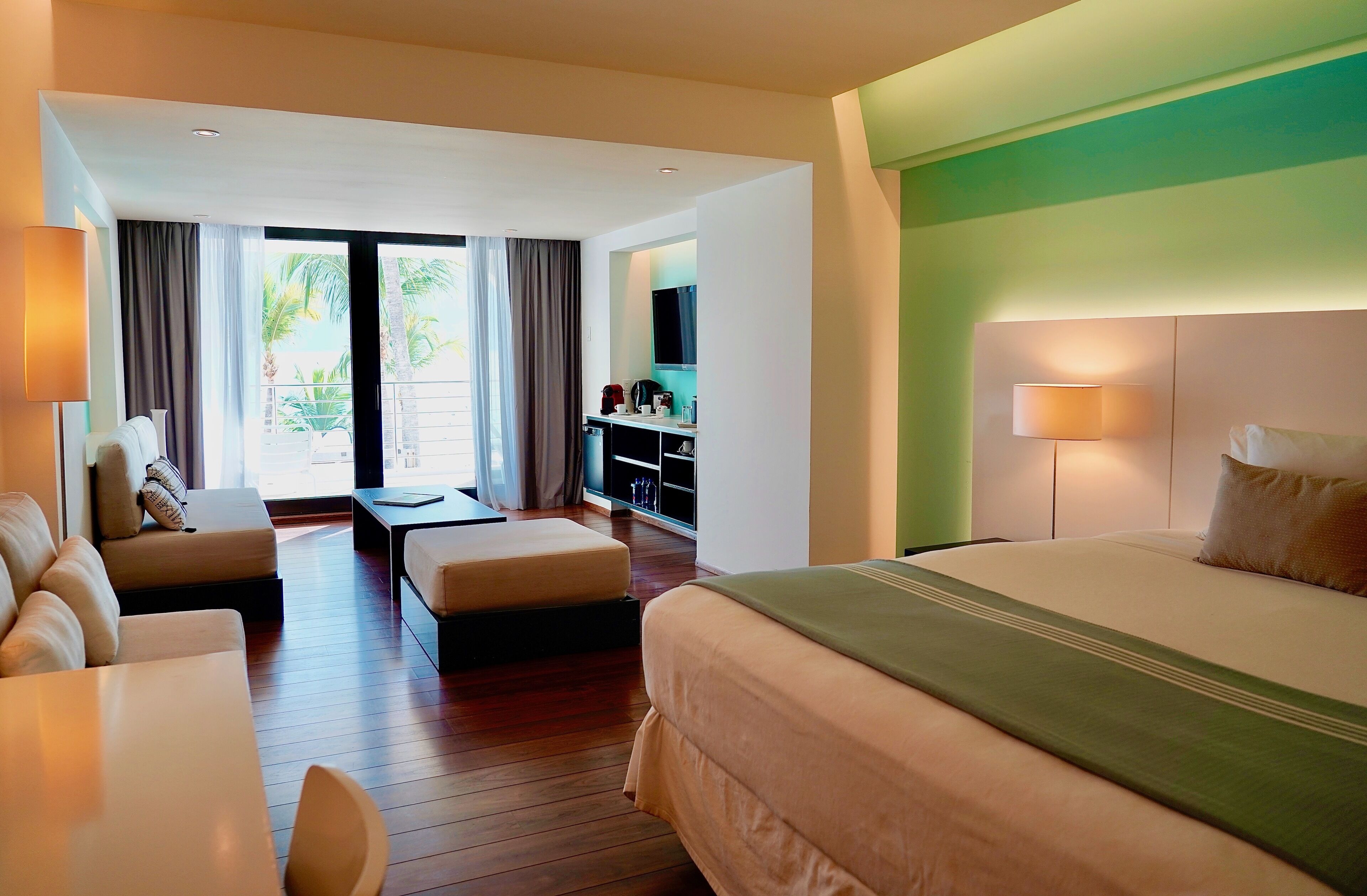 A hotel room with a large bed, a sofa, a dining table, a small kitchen area, and a view of palm trees.