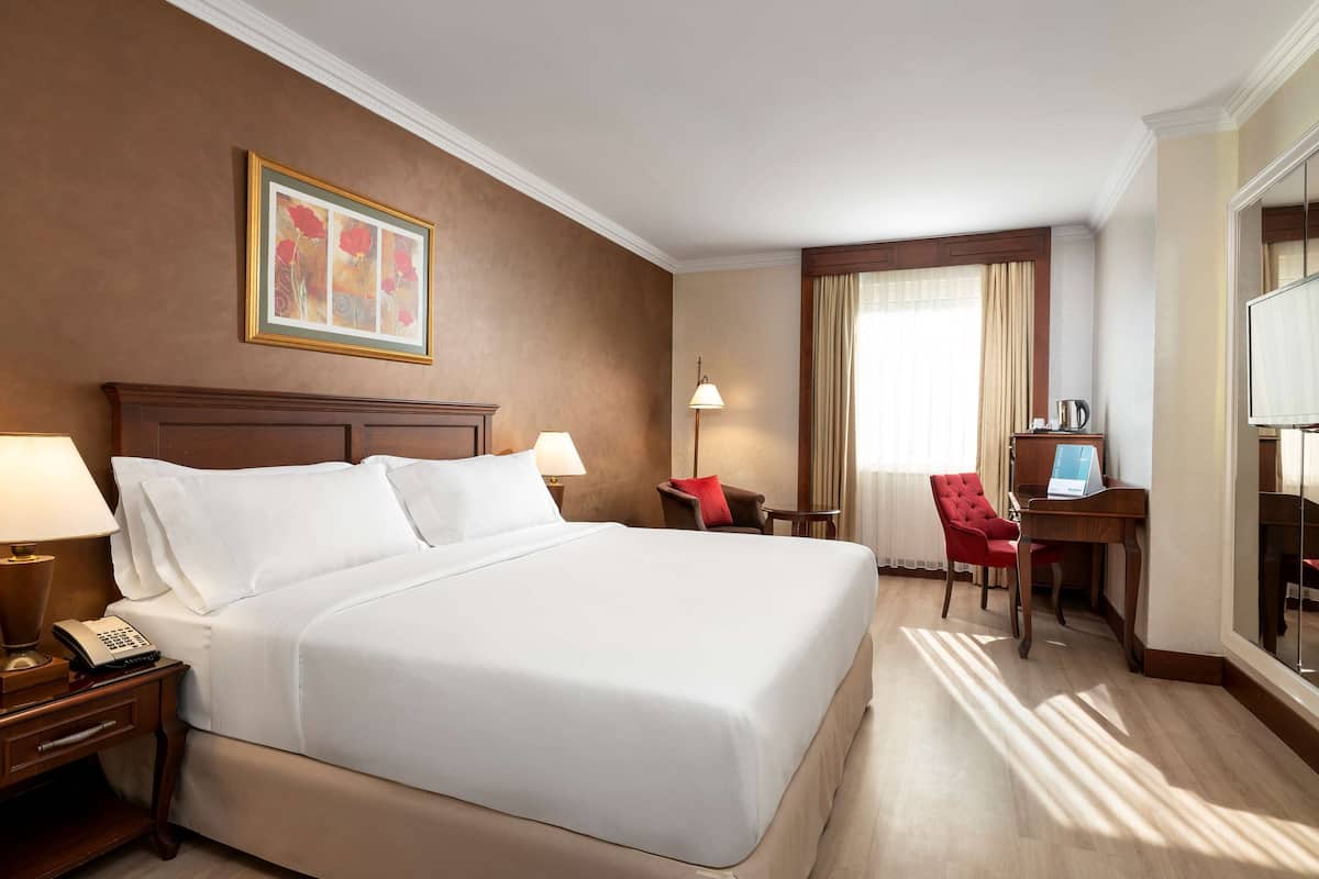 A hotel room with a large bed, a desk with a computer, a chair, a lamp, and a telephone on the nightstand.