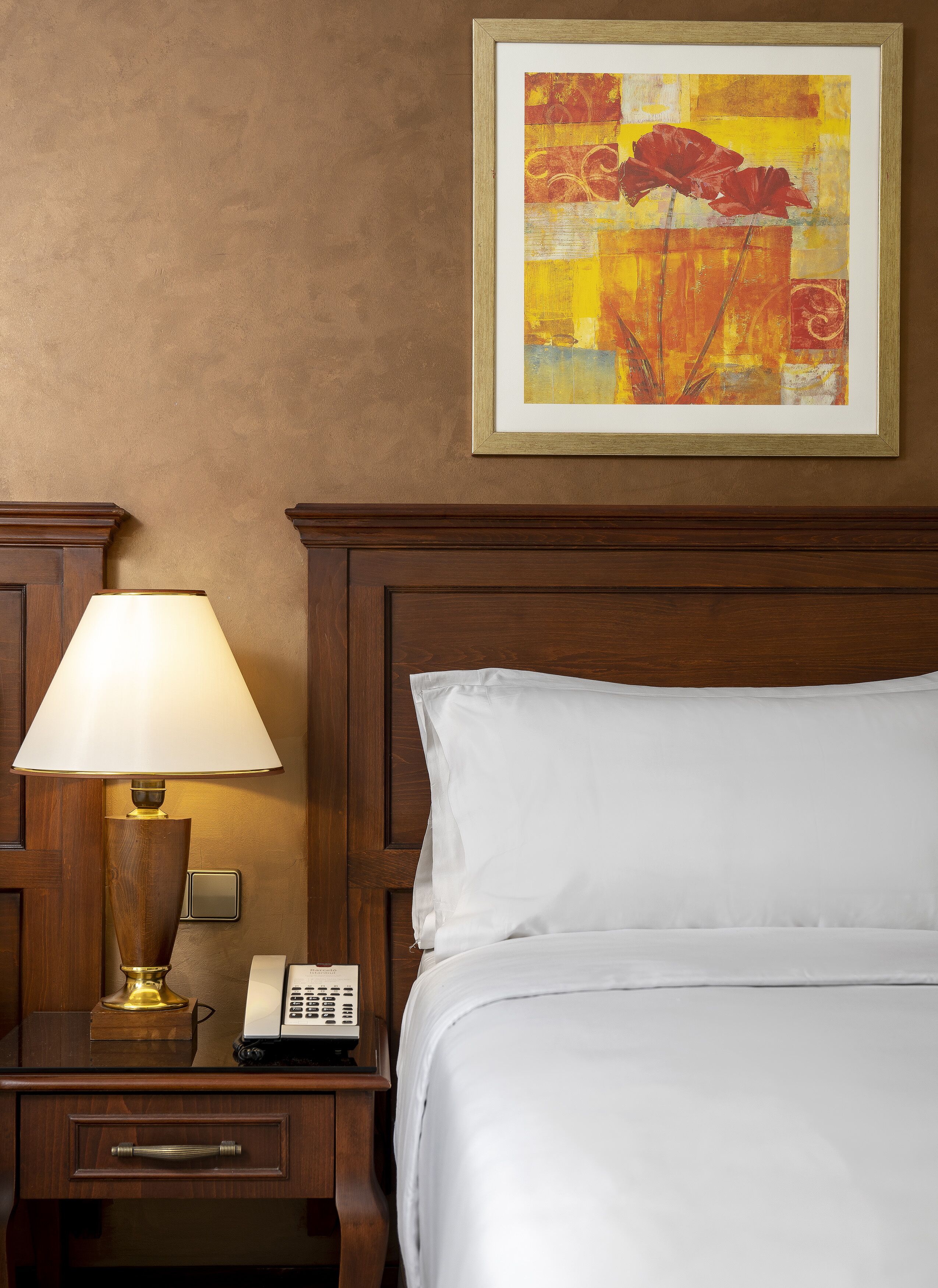 A hotel room with a bed, a nightstand with a lamp, a telephone, and a framed painting on the wall.