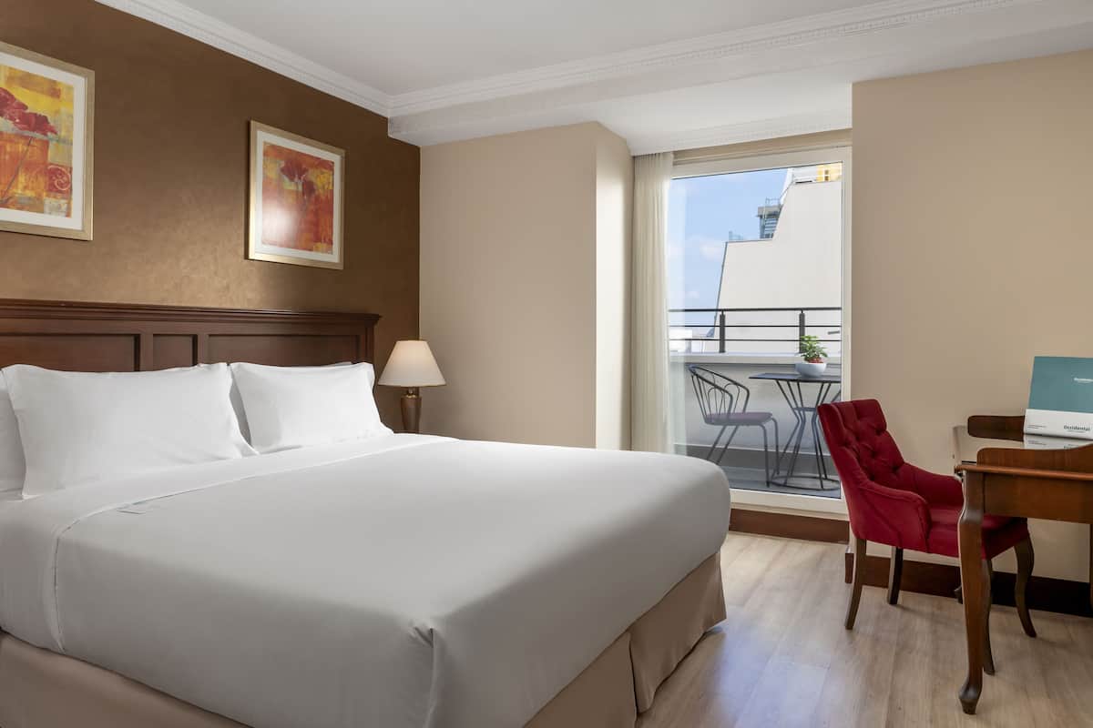 A hotel room with a large bed, a desk with a chair, a window with a view of the outside, and two framed pictures on the wall.