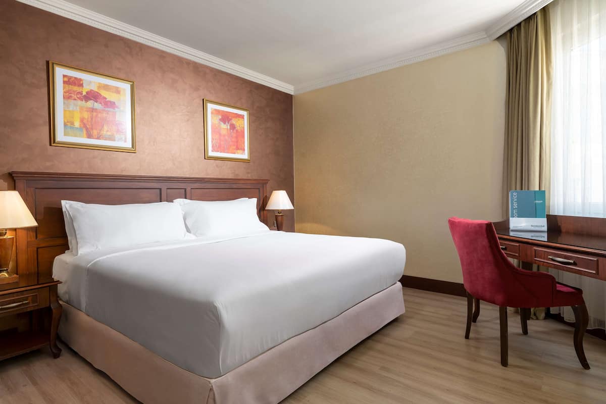 A hotel room with a large bed, two bedside lamps, a desk with a chair, and two framed artworks on the wall.