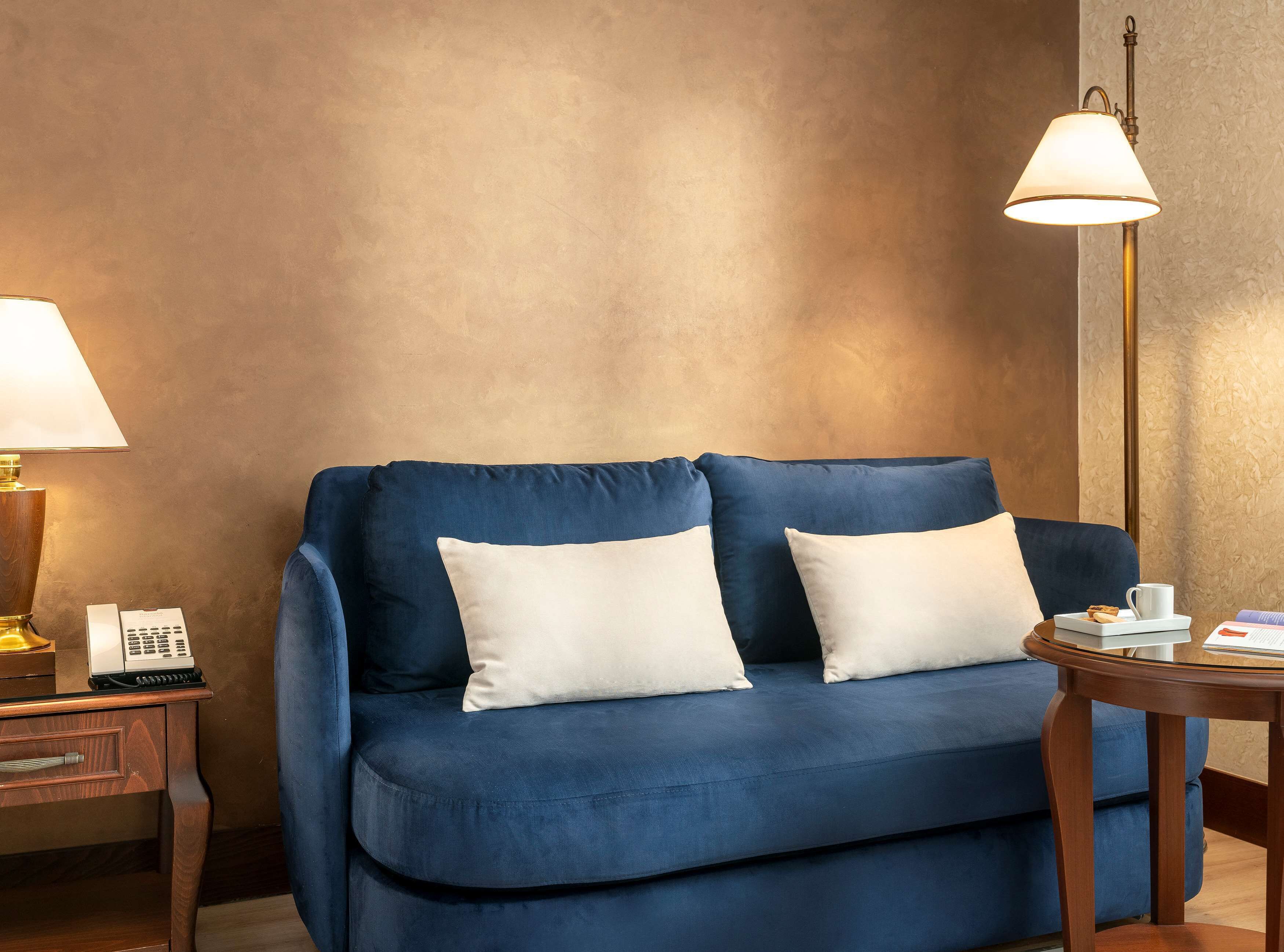 A blue sofa with two white cushions, a bedside table with a lamp, and a small table with a cup and a book.