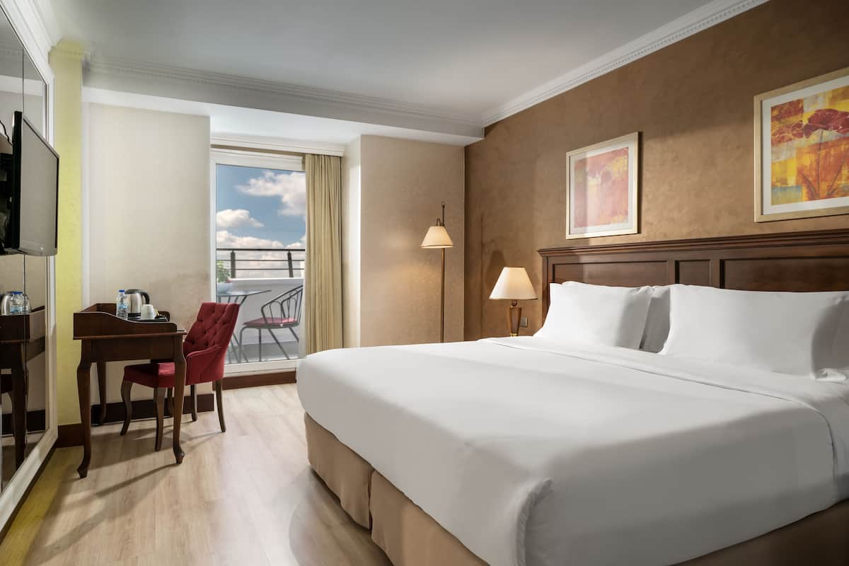 A hotel room with a large bed, a desk with a chair, a TV, and a balcony with a view of the sky.