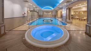 Una amplia piscina cubierta con un jacuzzi en el centro, rodeada de sillones de descanso y un techo con iluminación empotrada.