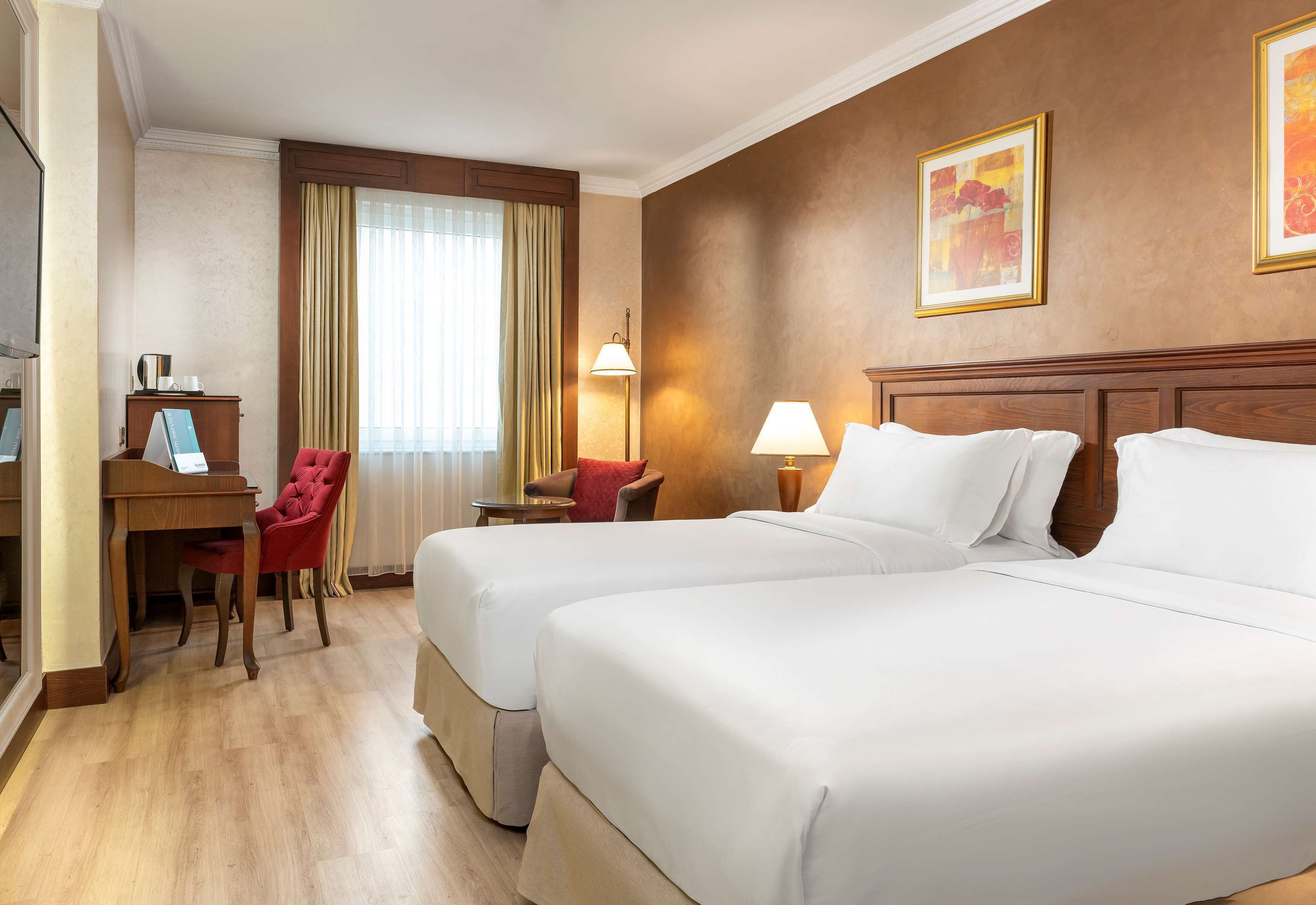 A hotel room with two beds, a desk, a chair, a lamp, and a window with curtains.