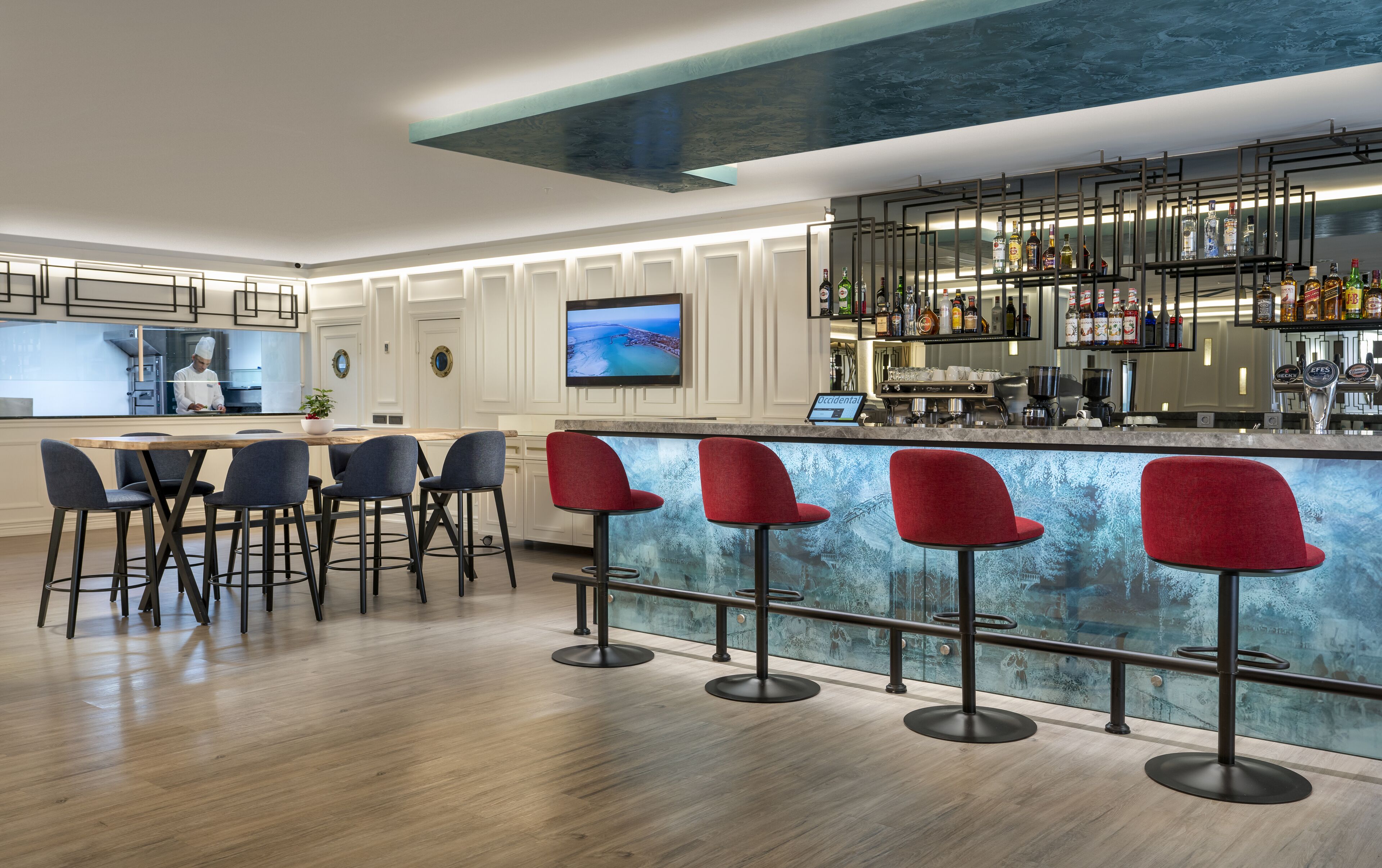 A modern bar area with a wooden bar counter, red bar stools, and a television screen displaying a scenic view.