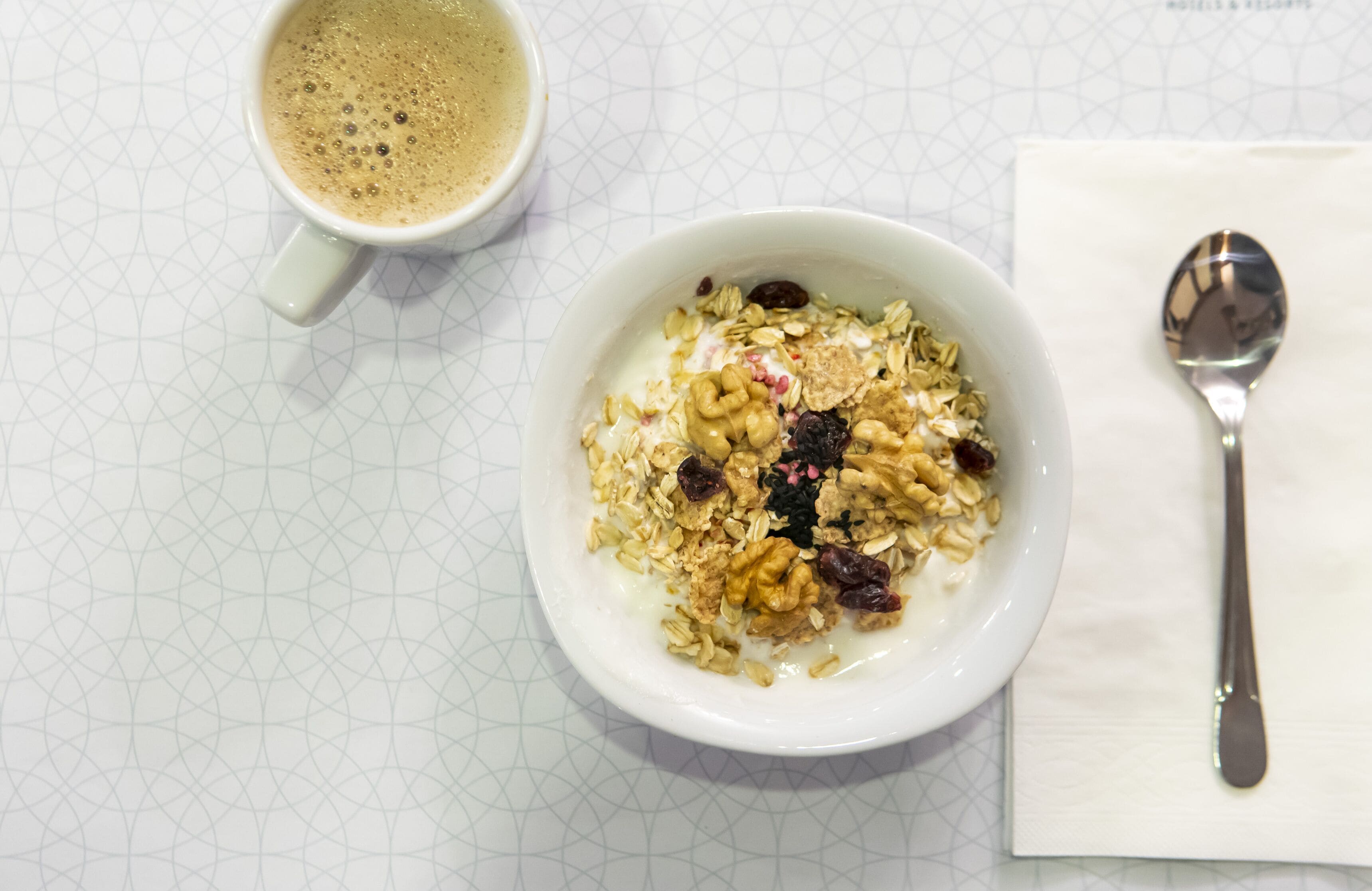A bowl of cereal with milk and a cup of coffee on a table with a geometric pattern.