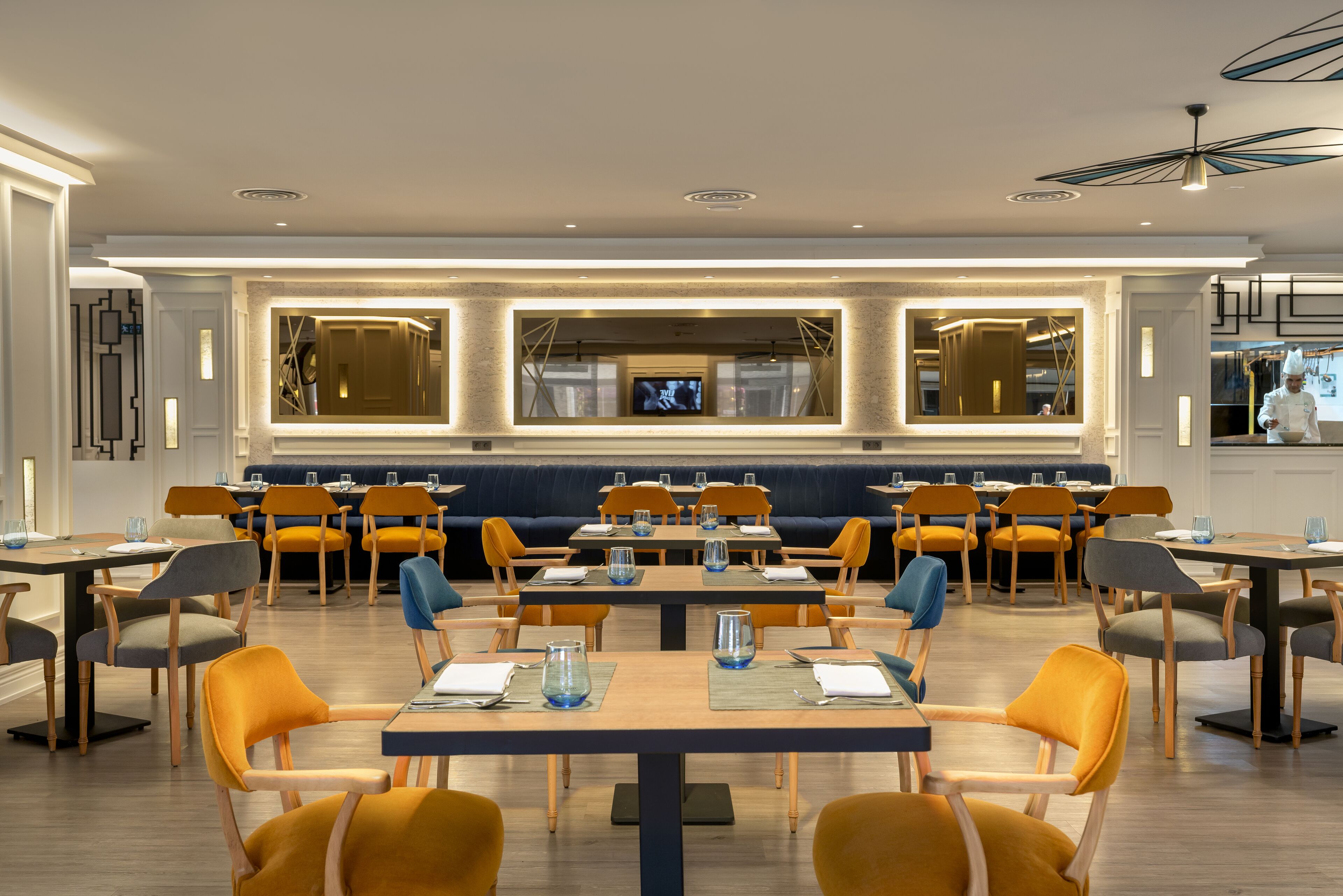 A modern dining area with multiple tables and chairs, a central seating area with a blue couch, and a television screen on the back wall.