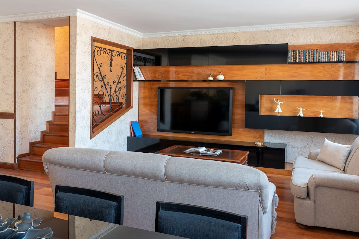 A modern living room with a flat-screen TV, built-in shelves, and a staircase.