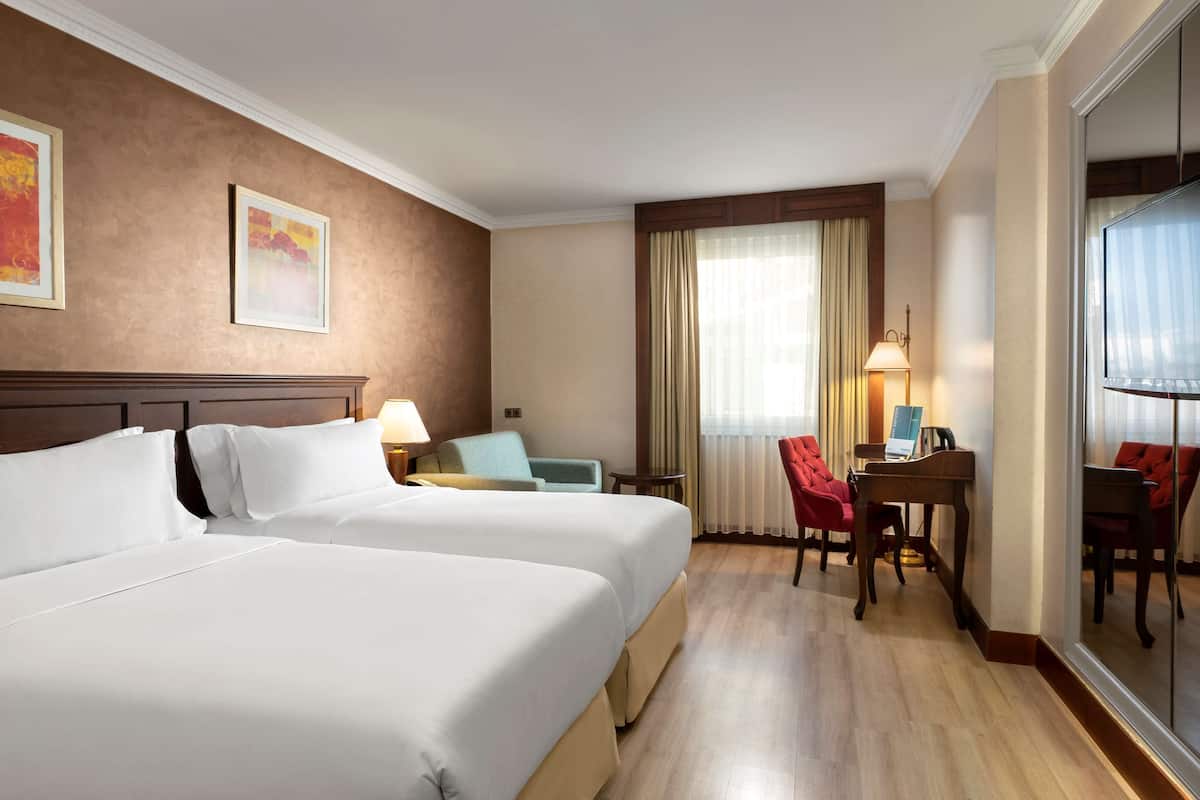 A hotel room with two beds, a desk with a piano, a chair, and a TV.