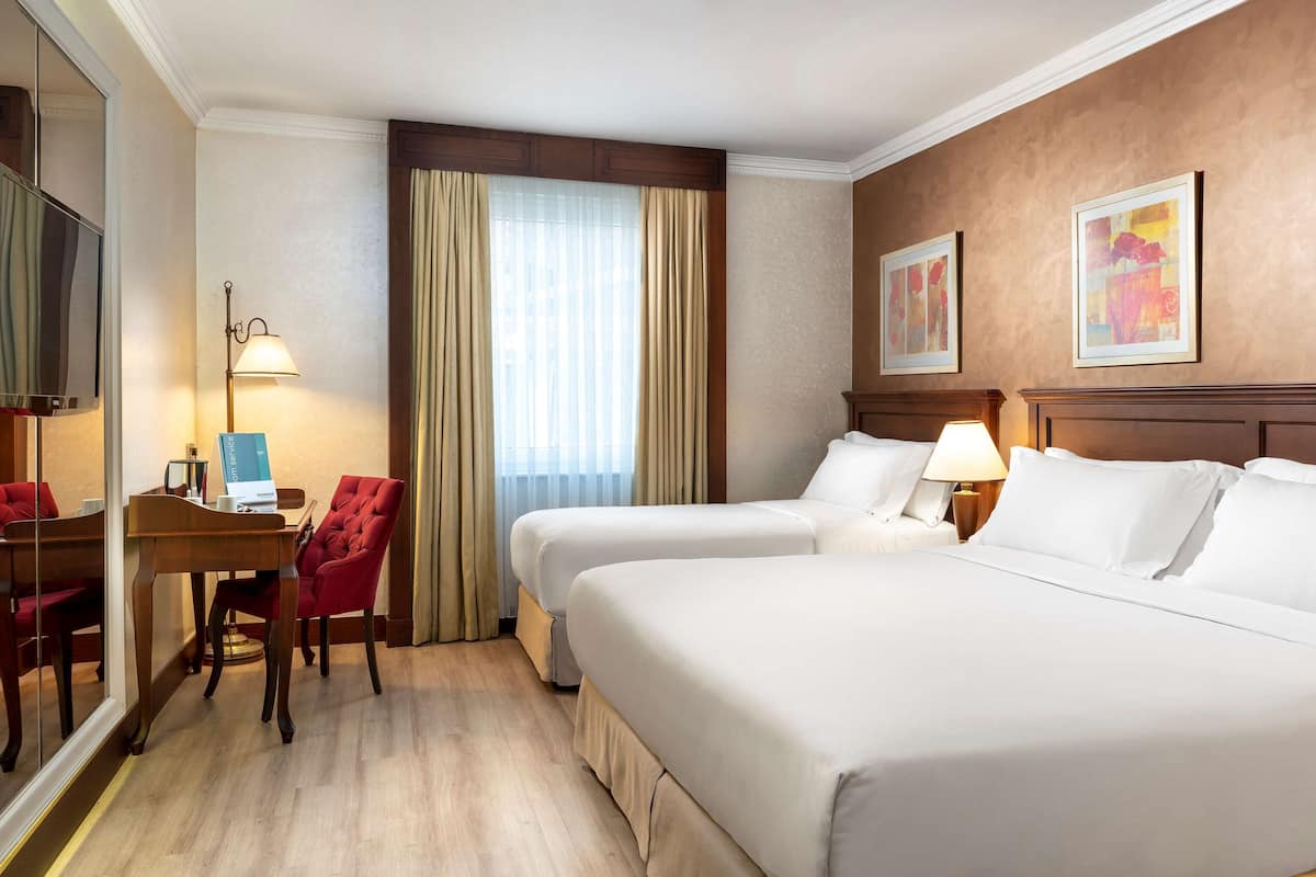 A hotel room with two beds, a desk with a chair, a TV, and a window with curtains.