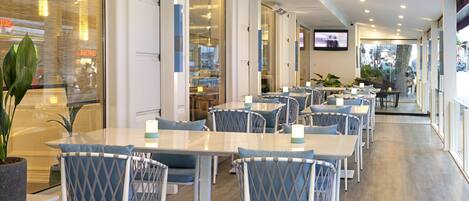 A modern restaurant with white tables and blue chairs, large windows, and a TV screen on the wall.