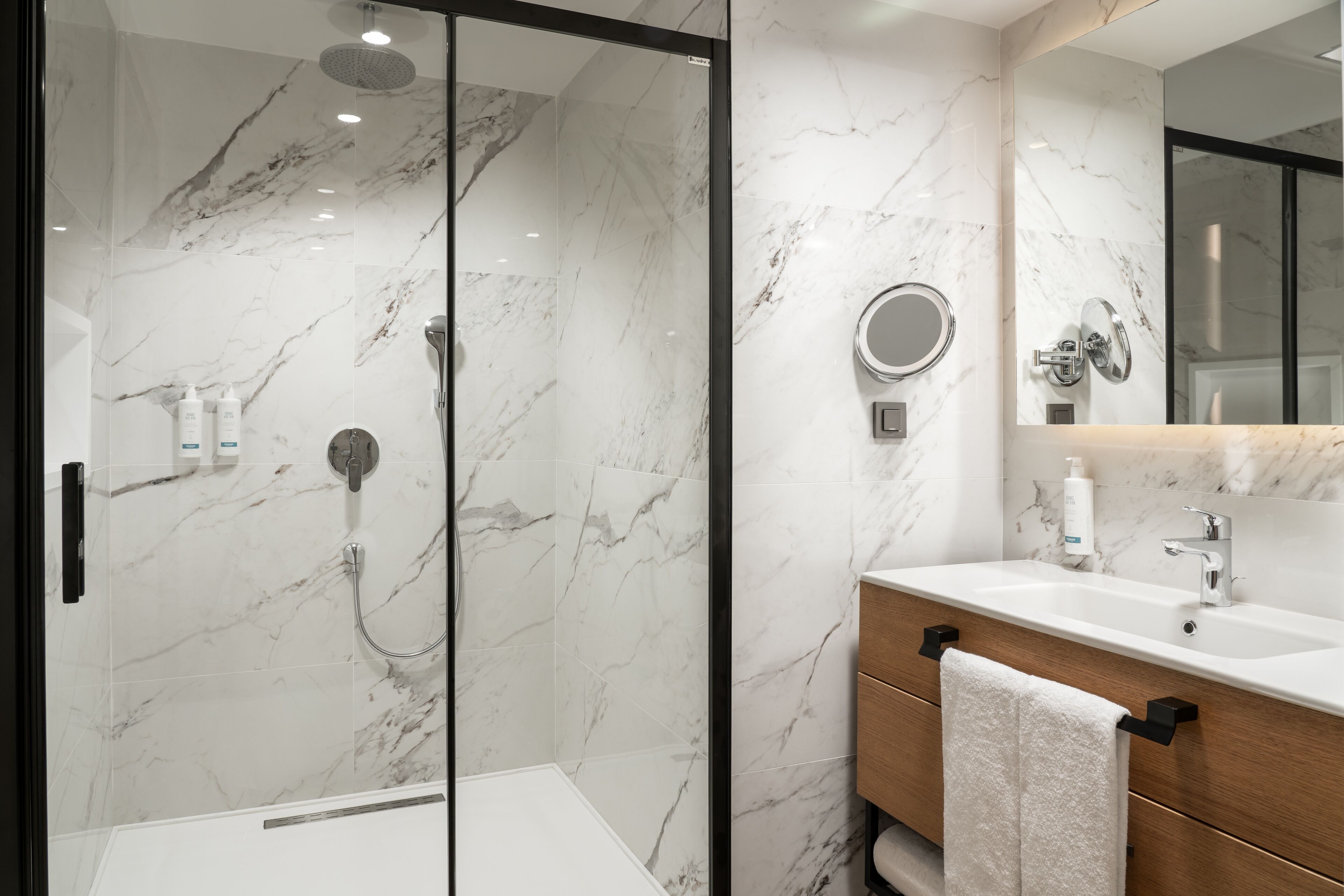 A modern bathroom with a glass-enclosed shower, a wooden vanity with a mirror, and marble walls.