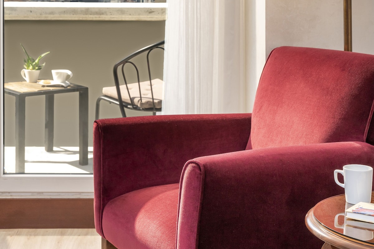 A red armchair by a window with a city view, a small table with a cup and saucer, and a floor lamp.