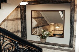 Una escalera de mármol con barandilla, una planta en maceta y un espejo grande que refleja una habitación con una lámpara en el techo.