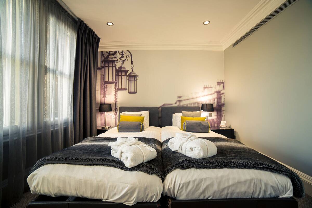 Ein Doppelbett mit zwei Kissen, ein Nachttisch mit Lampe und ein Fenster mit durchsichtigen Vorhängen.