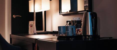A hotel room with a desk, a chair, a lamp, a coffee maker, and a mirror.