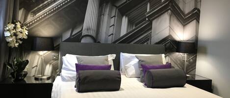 A neatly made bed with white bedding and multiple pillows, a black headboard, and a nightstand with a lamp and a potted plant.