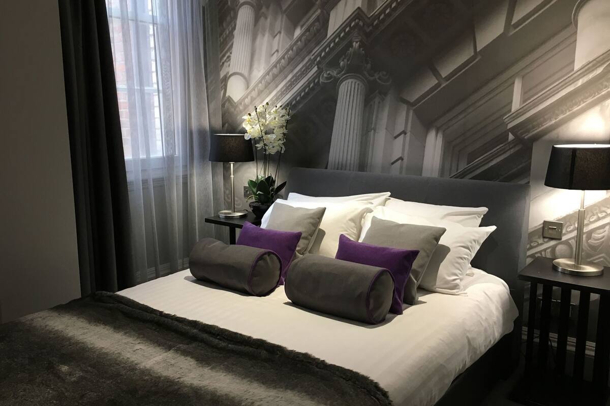 Ein modernes Hotelzimmer mit einem großen Bett, einem Nachttisch, einer Lampe und Blick auf ein klassizistisches Gebäude durch das Fenster.