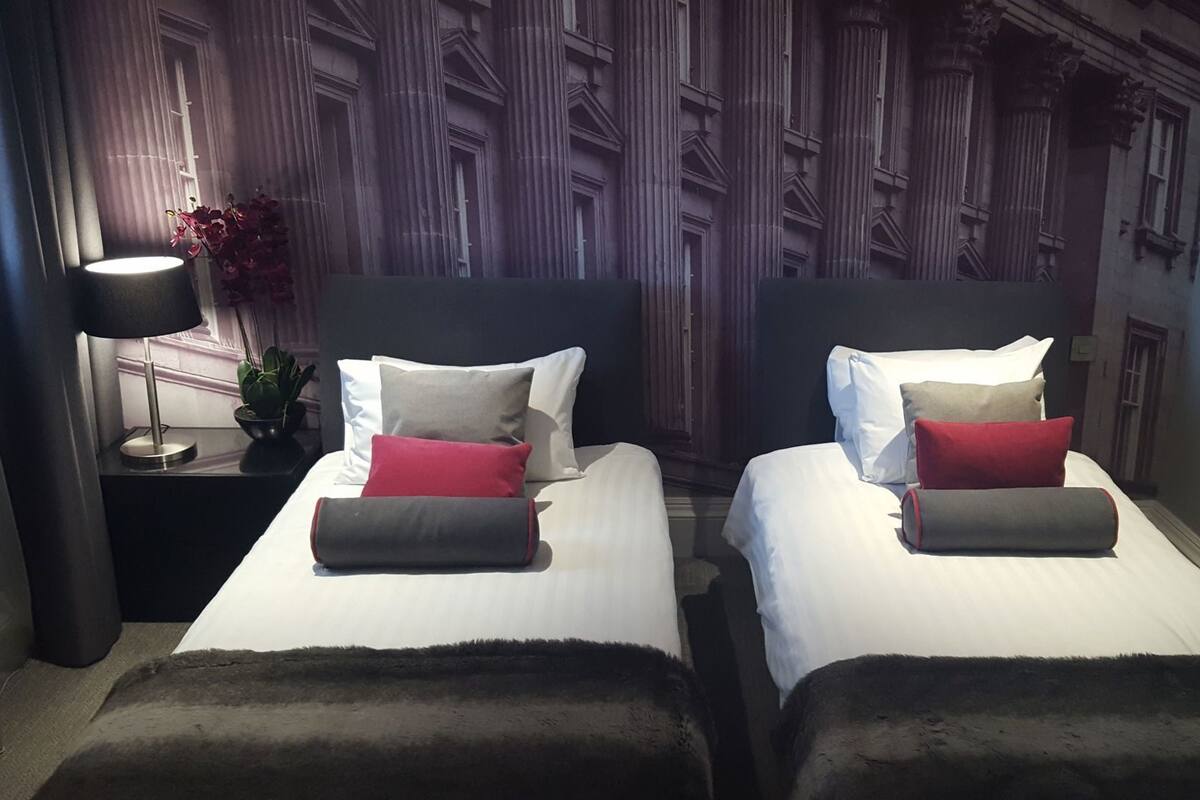 Ein Hotelzimmer mit zwei Betten, einem Nachttisch mit Lampe, einer Topfpflanze und einer Wanddekoration.