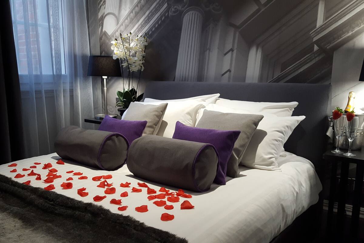 Ein Schlafzimmer mit einem großen Bett, einem Nachttisch, einer Lampe und Blick auf ein Gebäude durch das Fenster.