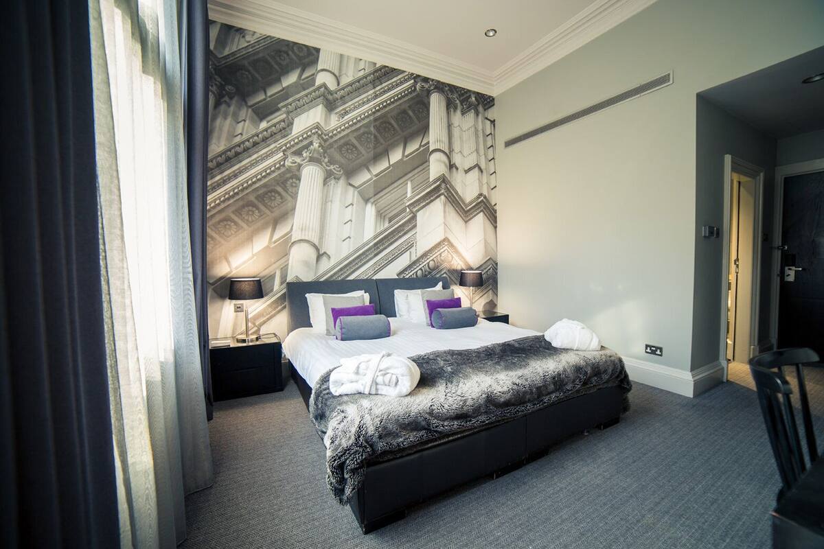 Ein Hotelzimmer mit einem großen Bett, einem Nachttisch, einer Lampe und Blick auf eine prächtige Treppe.