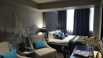 A modern hotel room with a large bed, a seating area with a chair and a small table, a lamp, and a decorative plant.