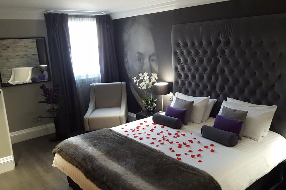 A hotel room with a large tufted headboard, a bed with white linens and red rose petals, a grey armchair, a side table with a lamp, and a mirror on the wall.