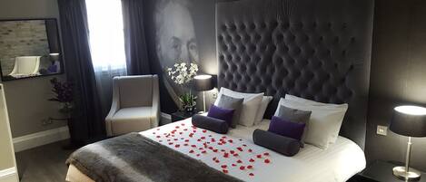 A hotel room with a large tufted headboard, a bed with white linens and red rose petals, a grey armchair, a side table with a lamp, and a mirror on the wall.