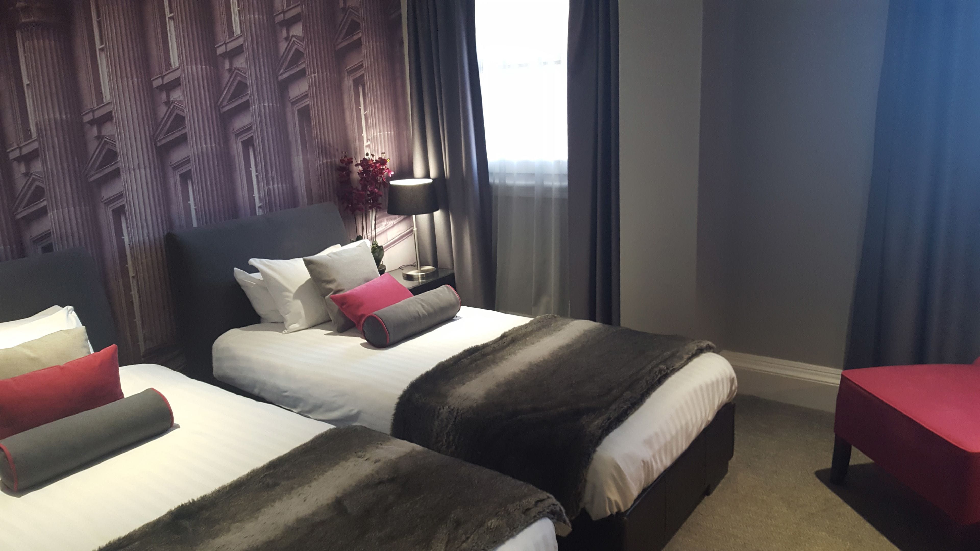 A hotel room with a large bed, a smaller bed, a nightstand with a lamp, a red chair, and a window with curtains.