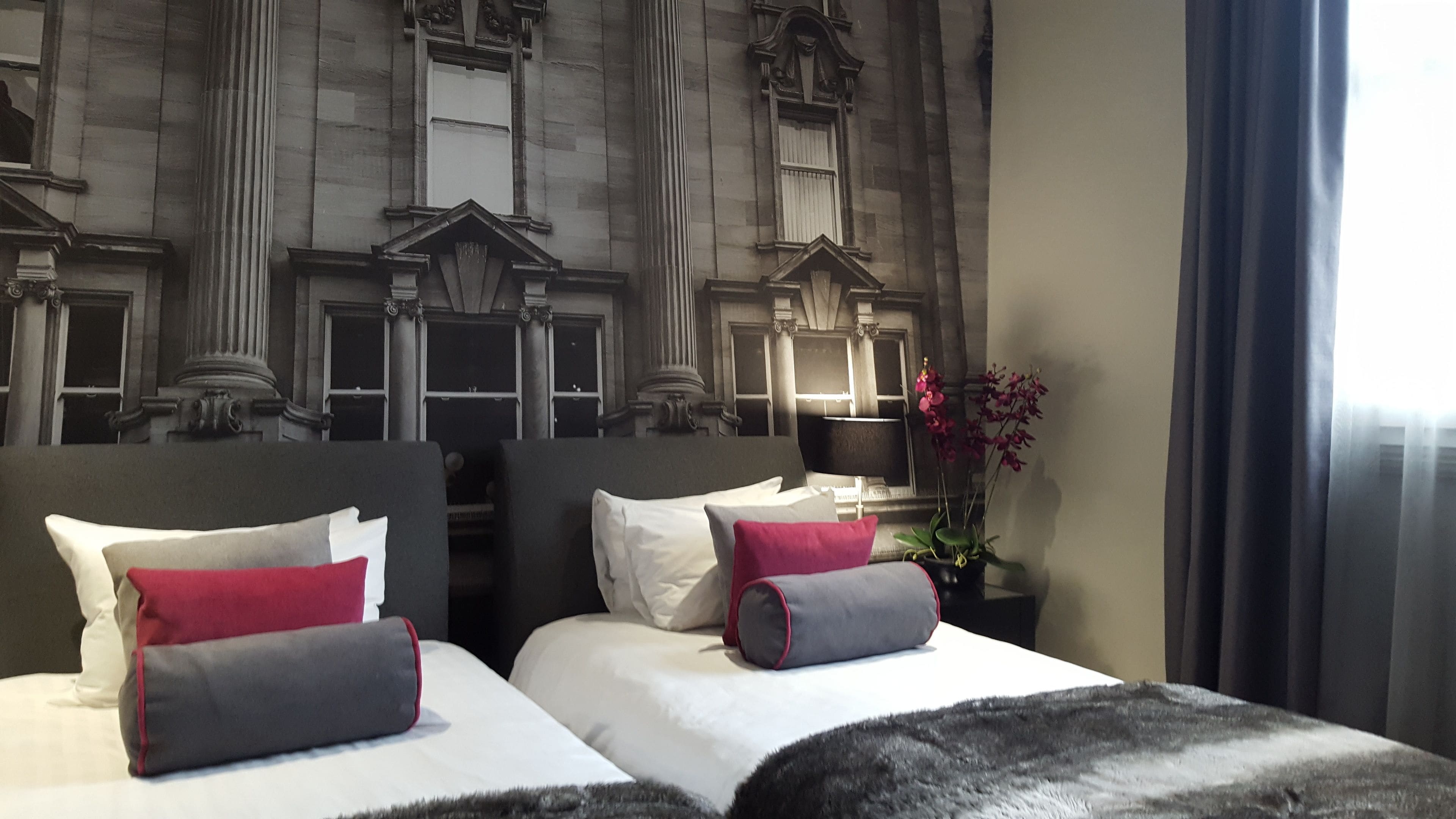 Habitación de hotel con dos camas, cada una con almohadas blancas y rojas, un ventanal con cortinas y una pared decorativa con arquitectura clásica.