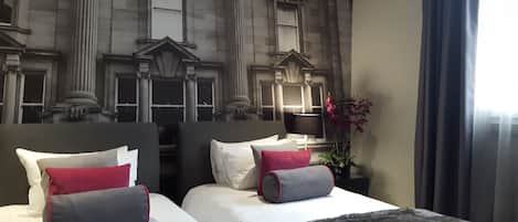 A hotel room with two beds, each with white and red pillows, a large window with curtains, and a decorative wall with classical architecture.