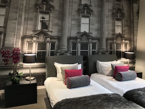 Quarto de hotel com duas camas, uma janela grande e um mural com fachada de edifício.