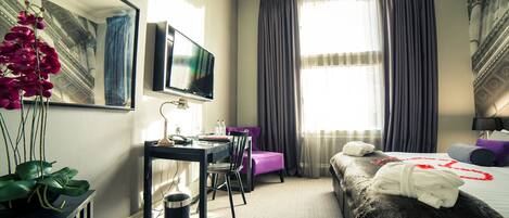 A hotel room with a bed, a desk with a chair, a TV, a window with curtains, and a plant.