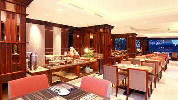 A hotel dining area with multiple tables and chairs, a buffet setup, and a large window with a view of the city.