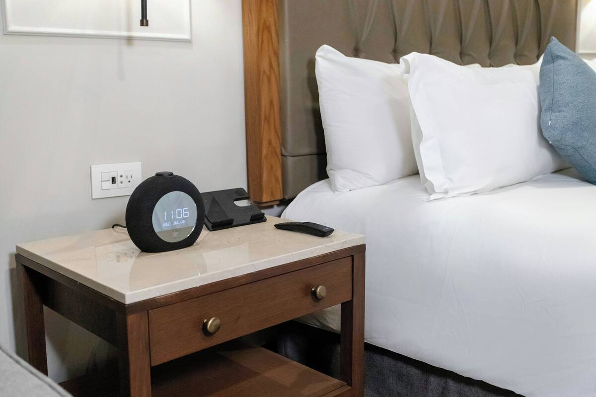 A hotel room with a bedside table, a digital clock, a remote control, and a bed with white and blue pillows.