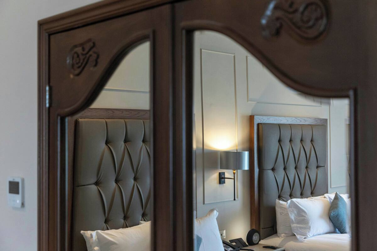 A bedroom with a tufted headboard, a bedside table with a lamp, and a mirror reflecting the room.
