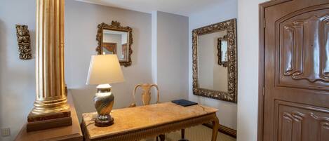 A room with a wooden table, a lamp, a chair, a mirror, and a door.