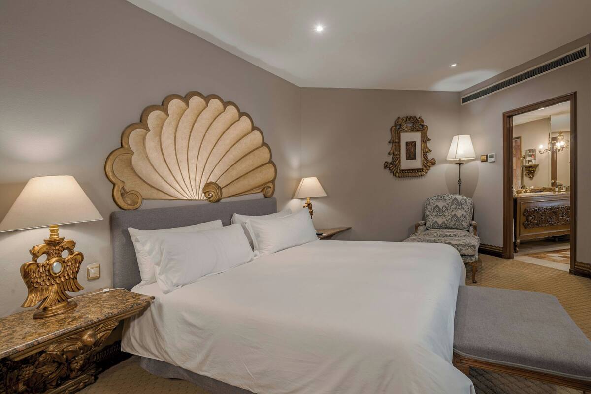 A hotel room with a large bed, two bedside tables with lamps, a chair, a mirror, and a decorative wall piece.