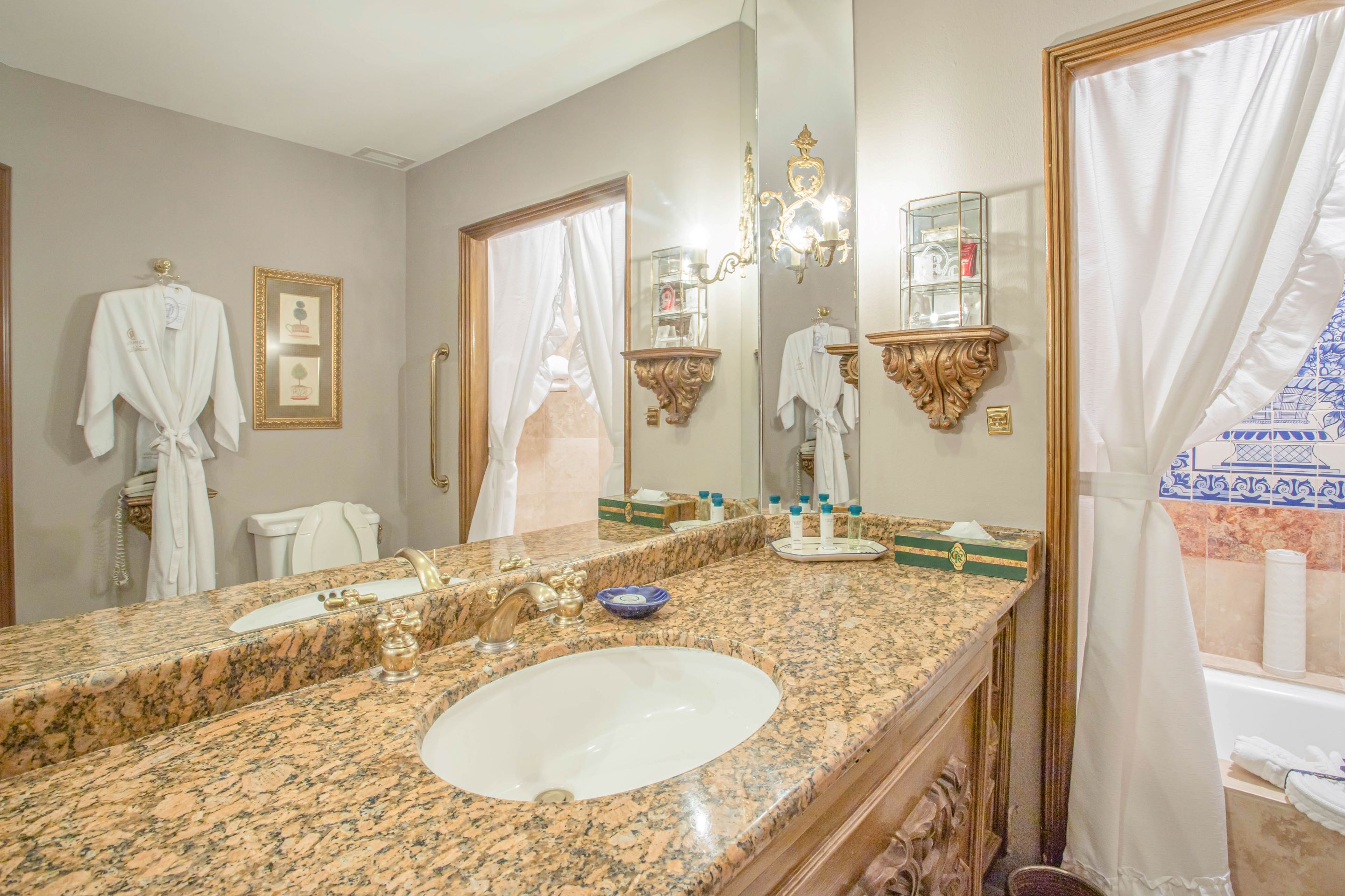 Un baño con encimera de granito, lavamanos, espejo grande y tocador con espejo y estantes.