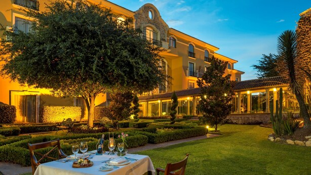 An outdoor dining area with a table set for two, surrounded by well-manicured hedges and a large tree. - Casa Mayor Saltillo, Hotel Hacienda (Saltillo)