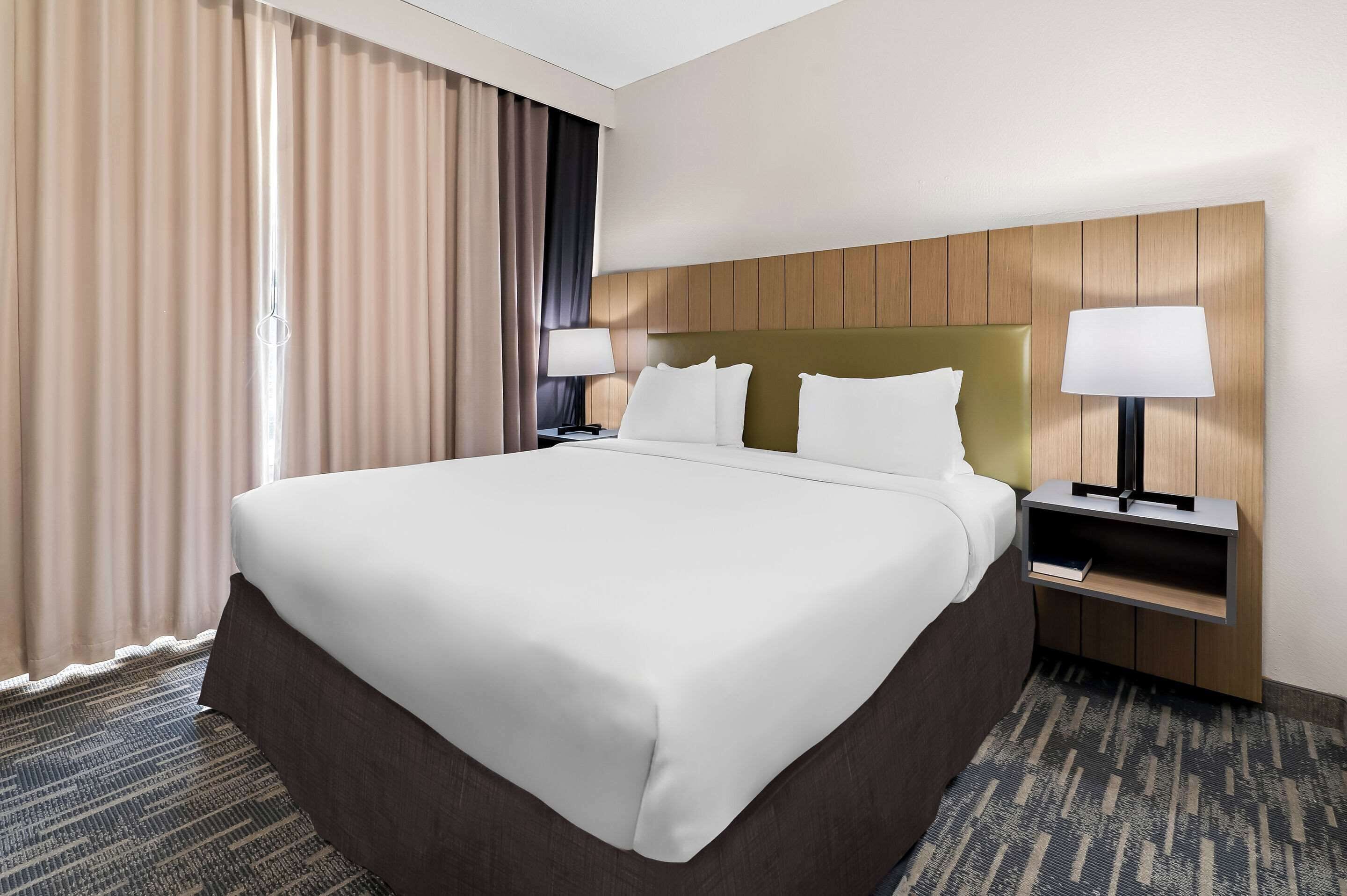 A hotel room with a large bed, two bedside tables with lamps, a headboard, and a view of the outside through curtains.