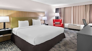 Quarto de hotel com uma cama grande, duas mesas de cabeceira com luminárias, uma cadeira vermelha, uma escrivaninha e uma janela grande com cortinas.