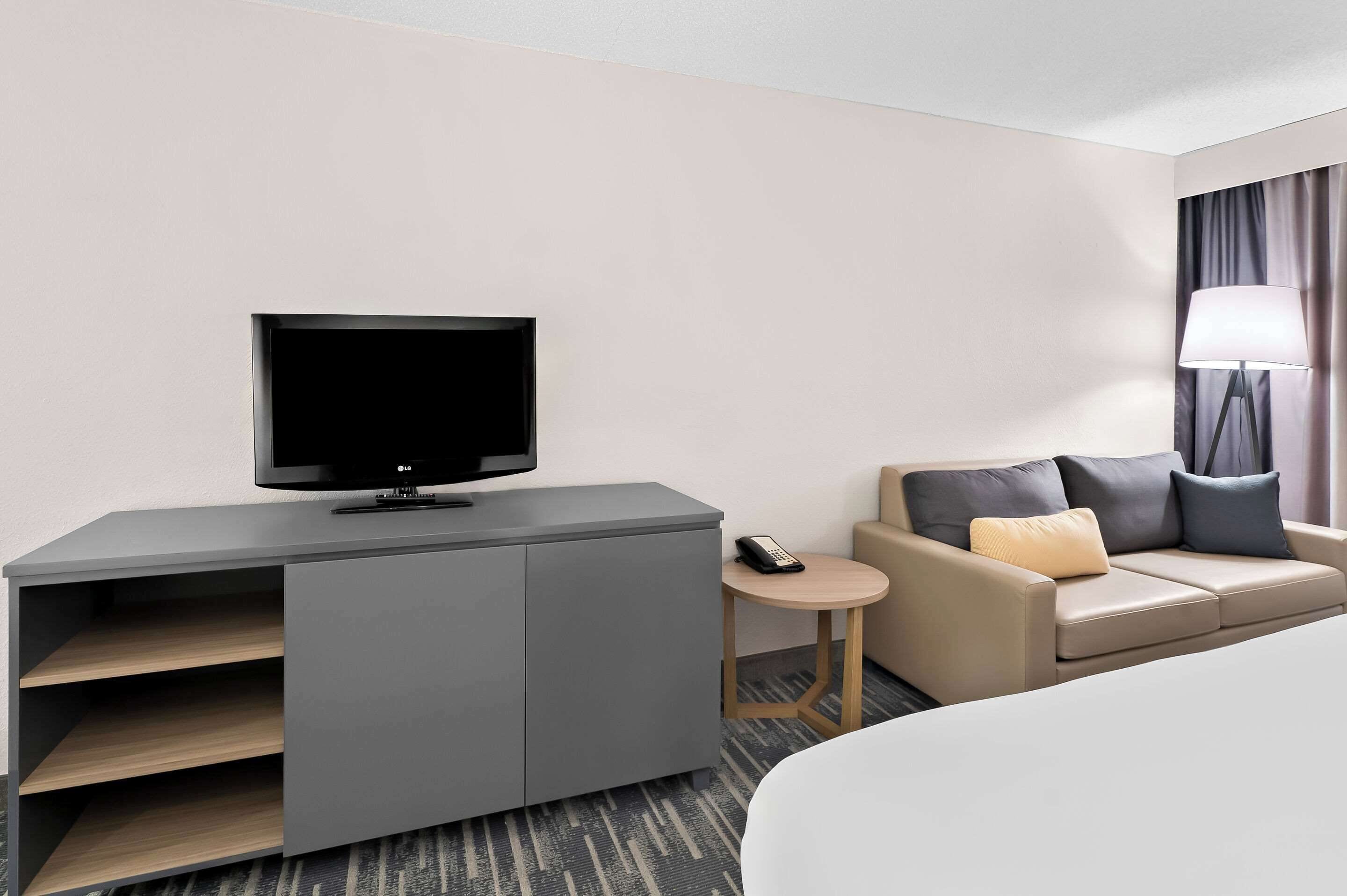 A hotel room with a flat-screen TV on a grey cabinet, a small round table with a remote, a beige sofa with cushions, and a lamp on a side table.
