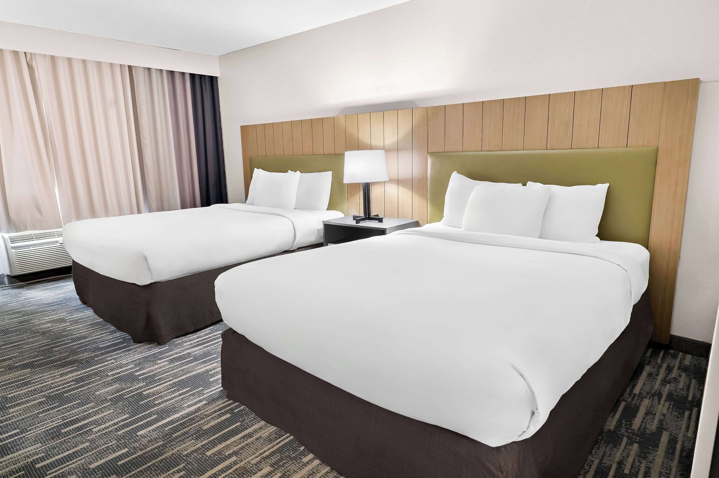 A hotel room with two beds, a nightstand, a lamp, and a large window with curtains.