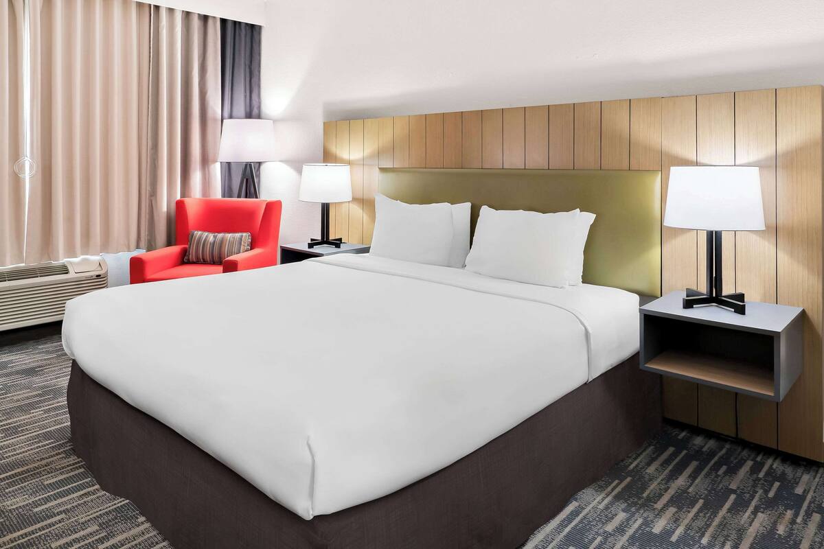 A hotel room with a large bed, two bedside tables with lamps, a red chair, and a wall-mounted shelf.