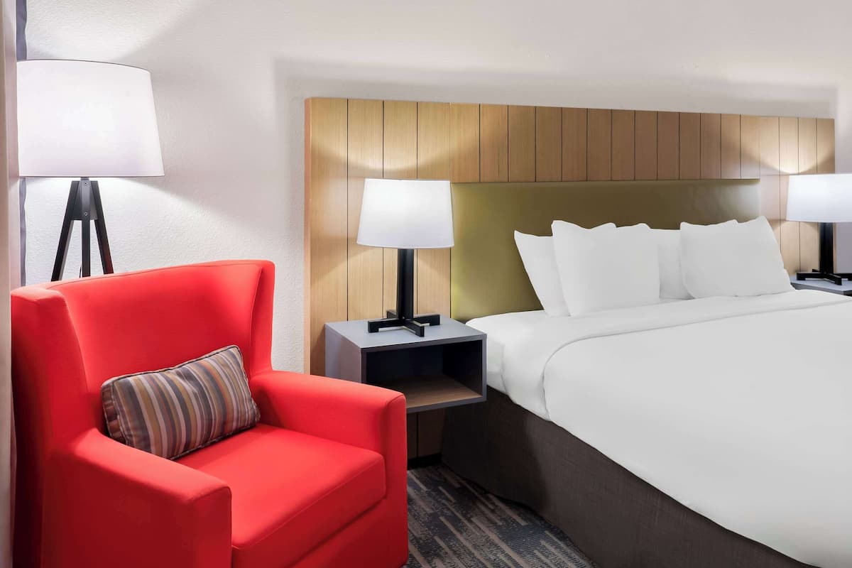 A hotel room with a large bed, two bedside tables with lamps, a red armchair, and a wooden headboard.