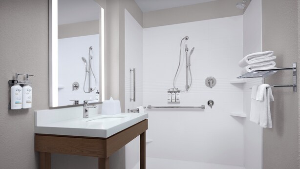 Un baño moderno con un espejo grande, un lavamanos blanco, una ducha con cabezal de ducha manual y toallas ordenadamente dobladas en un perchero.