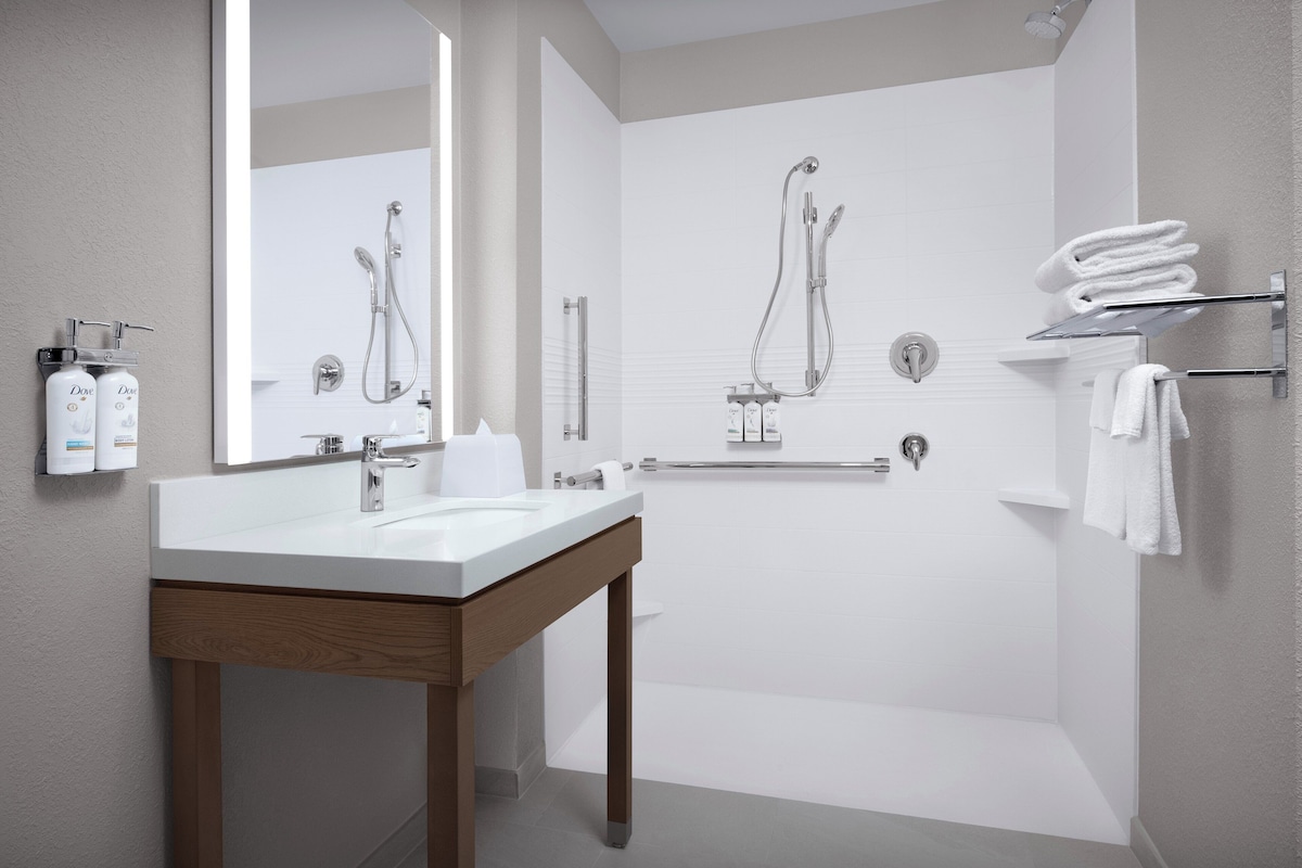 A modern bathroom with a large mirror, a white sink, a shower with a handheld showerhead, and neatly folded towels on a rack.