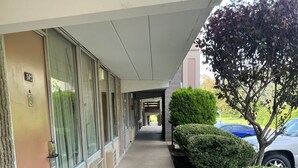 A hotel corridor with a covered walkway, a door marked '102', and a parking area with cars and greenery. - Travelers Inn (Dayton)